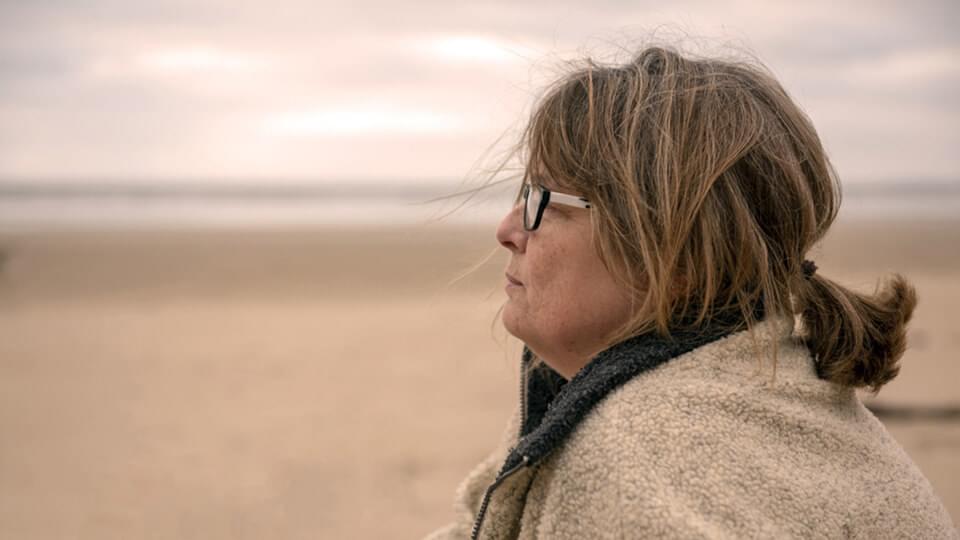 A middle aged lady sitting on a cold looking beach staring into the distance